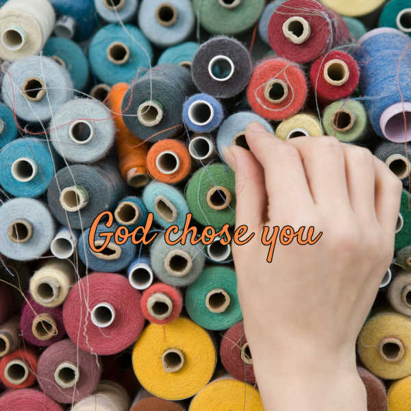 Photograph shows a hand selecting among densely arranged colorful yarn spools. Orange cursive text across center reads “God chose you,” while loose threads, cardboard cores, and varied red, blue, green, yellow, gray, and white fibers provide visual detail.
