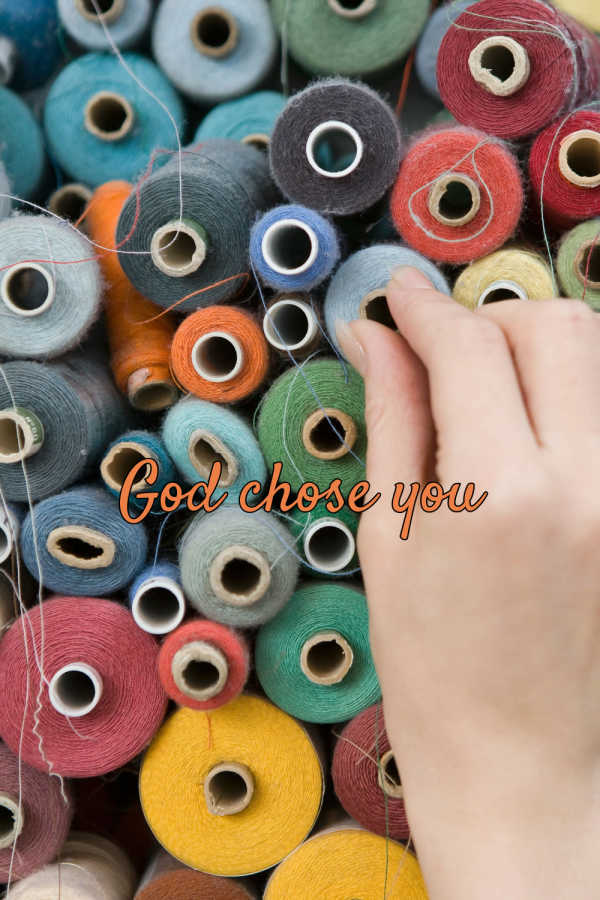 Photograph shows a hand selecting among densely arranged colorful yarn spools. Orange cursive text across center reads “God chose you,” while loose threads, cardboard cores, and varied red, blue, green, yellow, gray, and white fibers provide visual detail.