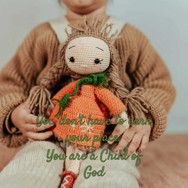 Photograph shows a child holding a handmade knitted doll, surrounded by warm muted colors and detailed yarn textures. Green script reads, “You don’t have to earn your place; you are a Child of God,”